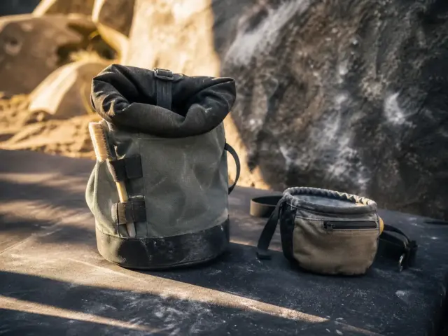 0-chalk-bag-bucket-hero Climber's chalk bucket and compact chalk bag sitting side by side on a crash pad at the base of a boulder problem