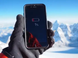 Everest summit phone life question: Smartphone showing low battery and cold warning held by gloved hand near Everest peak.