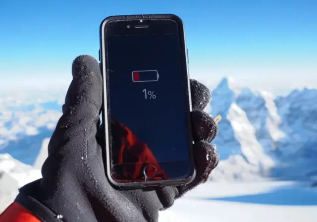 Everest summit phone life question: Smartphone showing low battery and cold warning held by gloved hand near Everest peak.