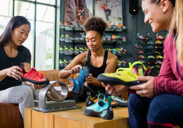Three diverse women carefully selecting and trying on different models of women's climbing shoes in a well-stocked gear shop, illustrating the ultimate guide to fit and performance.