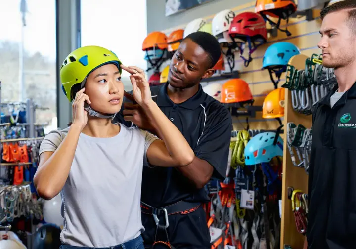 Diverse climbers in a gear shop, one trying on a climbing helmet while the other or a store employee advises, illustrating key factors for choosing the right helmet as their shield.