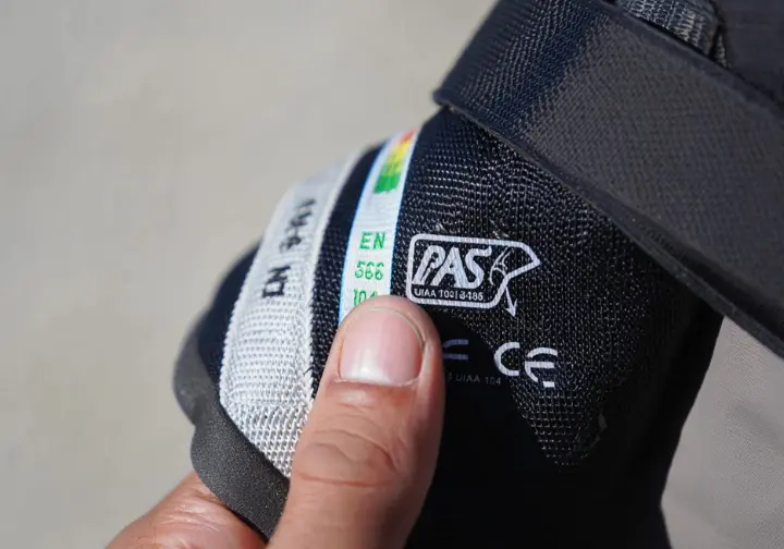 Extreme close-up of UIAA and CE safety certification labels on a climbing PAS, with a finger pointing to them, highlighting the importance of standards for anchor chain components.