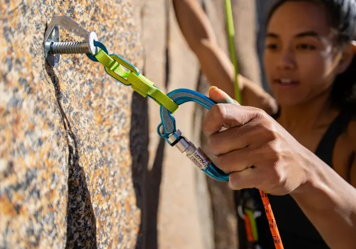Climber's gloved hands securely connecting a chain-style PAS to a fixed rock anchor with a fully locked carabiner, demonstrating proper anchor chain technique.
