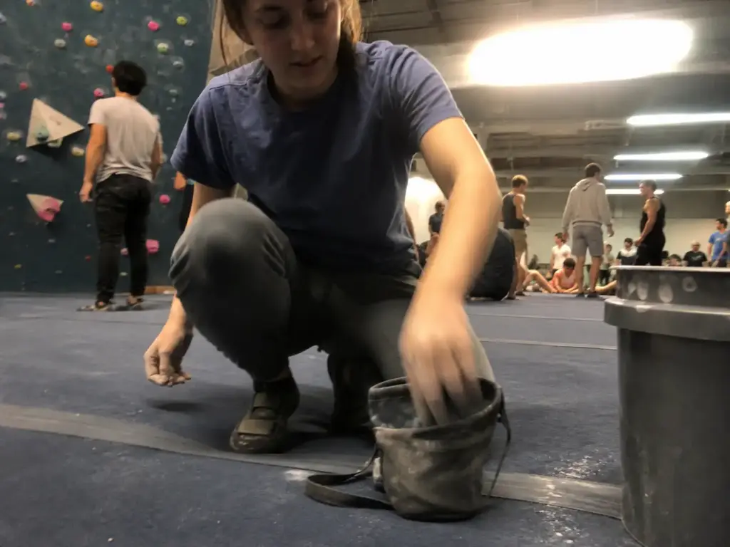Climber crouching to pick up a compact chalk bag while a larger chalk bucket sits open on the gym floor beside them