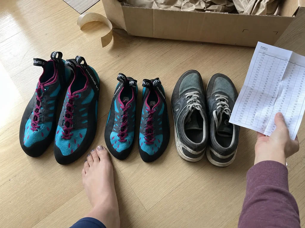 Two sizes of La Sportiva Tarantulace women's climbing shoes laid on the floor next to a street sneaker
