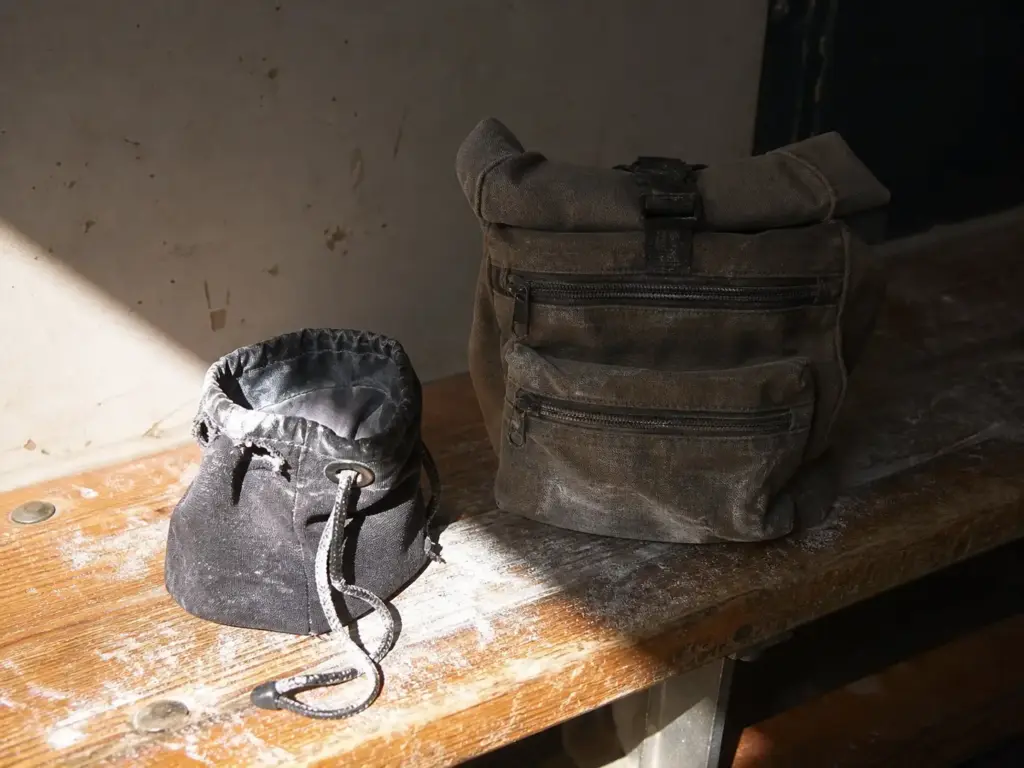 A worn budget chalk bag with a frayed drawstring next to a heavier canvas bag with reinforced stitching on a bench