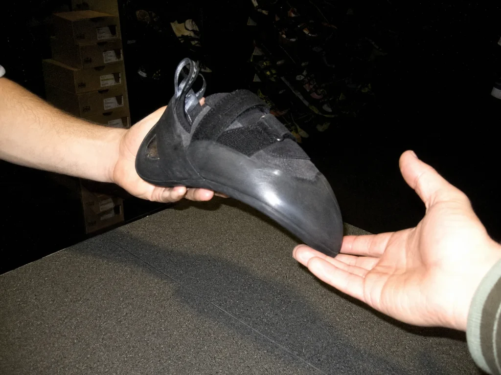 Gym employee handing an aggressive downturned climbing shoe across a dim fitting counter