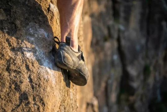 Best Climbing Shoes Tested: Performance & Fit Guide Close up of a climbing shoe heel hook on sandstone rock showing rubber deformation and calf muscle tension.