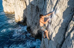 Damian Cook: The DWS Climber’s Life & Safety Lessons A climber deep water soloing on a steep limestone cliff above the ocean, representing the sport Damian Cook pioneered.
