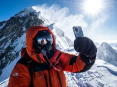 Phones in the High-Altitude Zone: A Summit Survival Protocol A climber in a full down suit on the Everest ridge holding a frosted smartphone against a backdrop of deep blue sky and blowing snow.