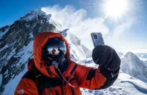 Phones in the Death Zone: A Summit Survival Protocol A climber in a full down suit on the Everest ridge holding a frosted smartphone against a backdrop of deep blue sky and blowing snow.