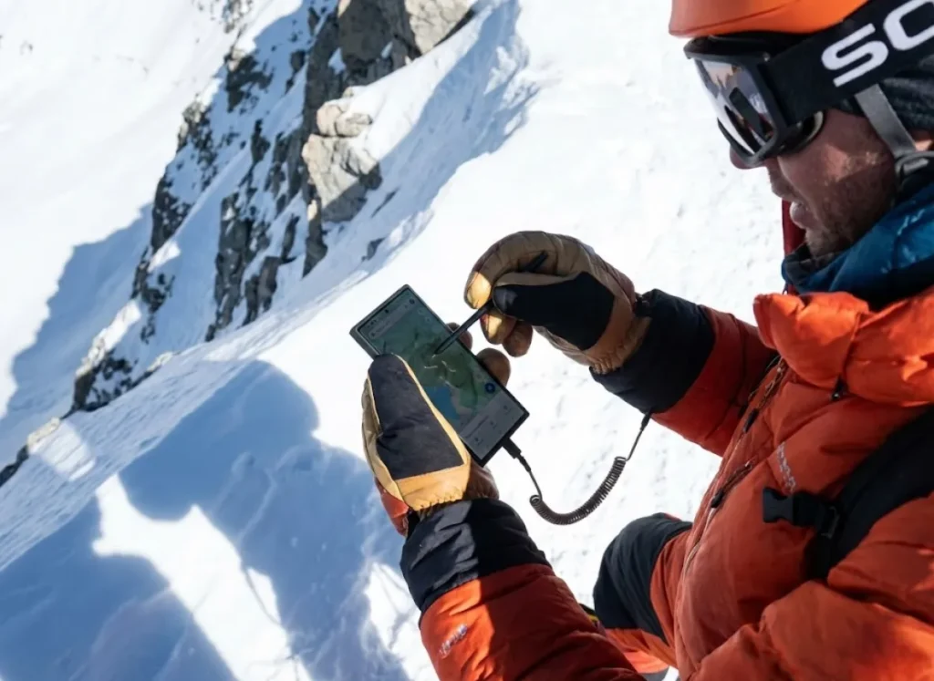 A mountaineer wearing thick mittens using a stylus pen to operate a smartphone screen on a snowy peak.
