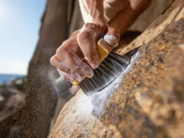 Climber's hand using a boar's hair rock climbing brush to clean chalk and debris off a rock hold, achieving max grip for better climbing performance.