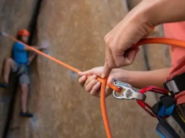 Close-up of a skilled belayer managing a rope through a belay device, with a climber in the background, illustrating how to perfect your belay in climbing for safety and skills.