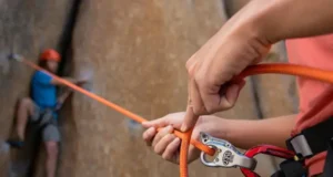 Perfect Your Belay in Climbing: Safety & Skills Close-up of a skilled belayer managing a rope through a belay device, with a climber in the background, illustrating how to perfect your belay in climbing for safety and skills.