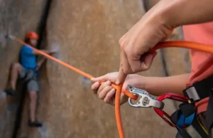 Perfect Your Belay in Climbing: Safety & Skills Close-up of a skilled belayer managing a rope through a belay device, with a climber in the background, illustrating how to perfect your belay in climbing for safety and skills.