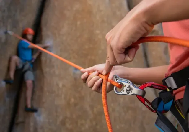 Close-up of a skilled belayer managing a rope through a belay device, with a climber in the background, illustrating how to perfect your belay in climbing for safety and skills.