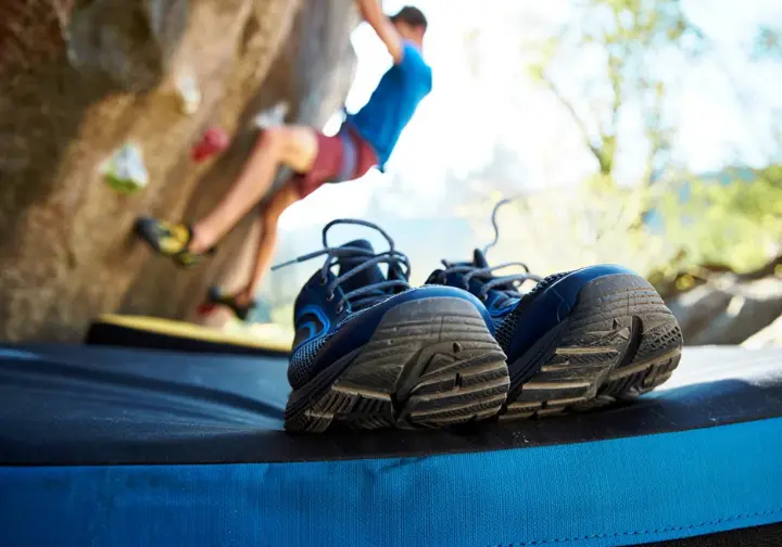Athletic sneakers placed on the sidelines of a climbing gym or crag, with a climber using proper climbing shoes in the background, showing why sneakers don't work for climbing.
