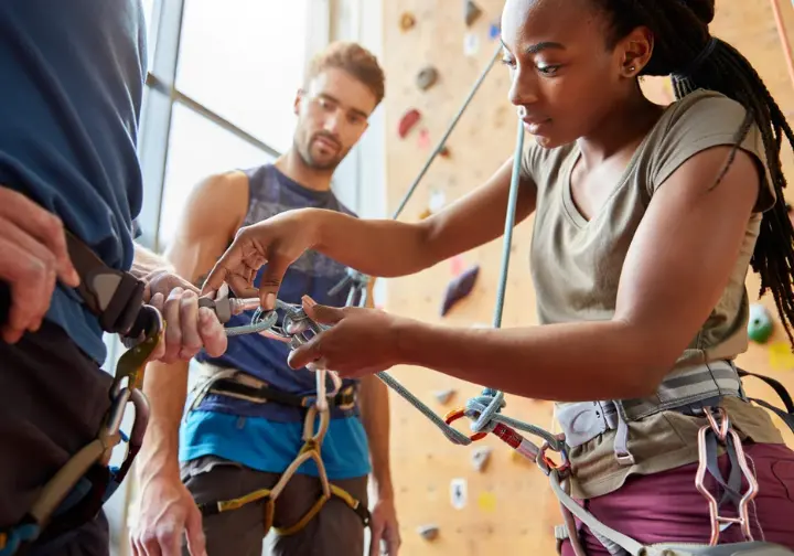 Two diverse climbers conducting a thorough pre-climb partner check (harness, knot, belay device, carabiner), highlighting core belay safety protocols for every climb.