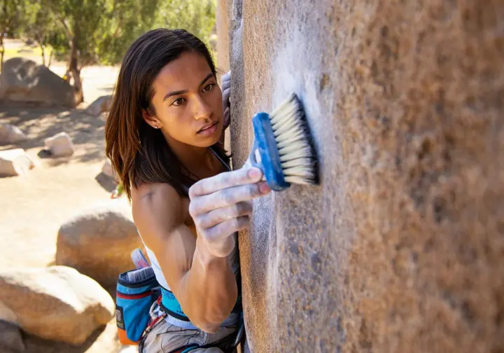 Climber demonstrating effective technique with a rock climbing brush on a specific hold outdoors, with another brush type nearby, illustrating how to choose and use brushes.