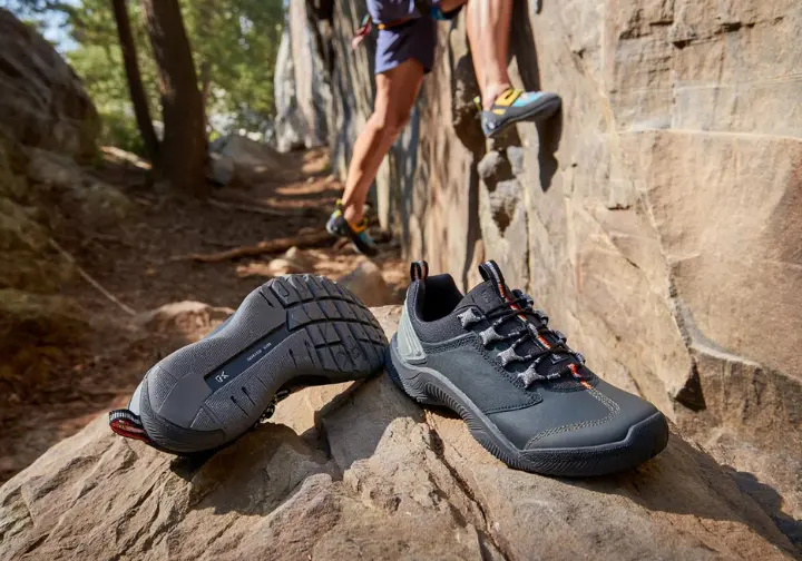 A pair of technical approach shoes at the base of a crag, with a climber in climbing shoes in the background, defining the approach shoe as the climber's "other sneaker".