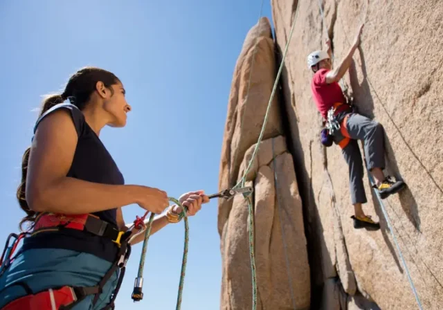Female belayer expertly managing rope for a male climber on a sunlit rock face, illustrating the ultimate answer to how to belay in climbing.