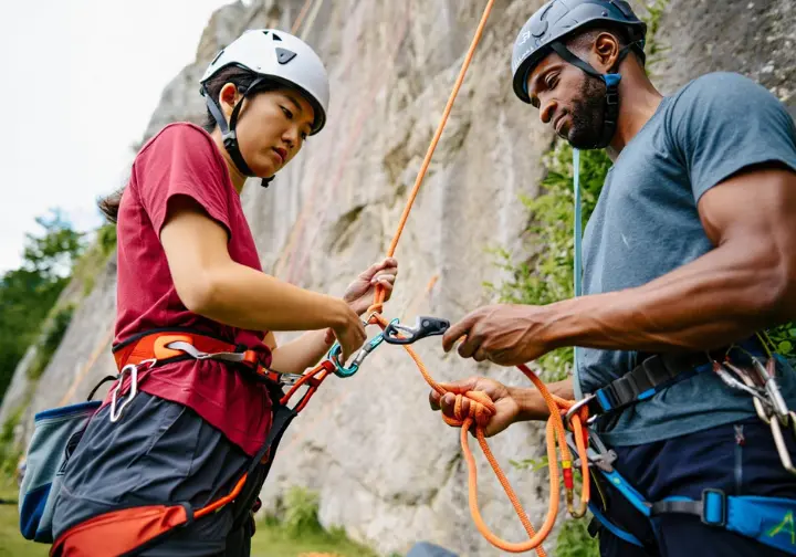 Two diverse climbers performing indispensable pre-climb safety checks (partner check) on harness and belay device, crucial for learning how to belay.