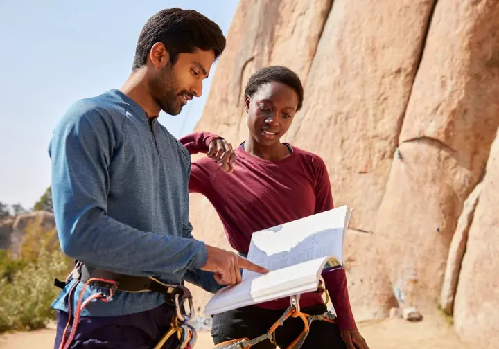 Climber and belayer discussing top-rope vs lead climbing options from a guidebook at a crag, learning how to belay in different scenarios.