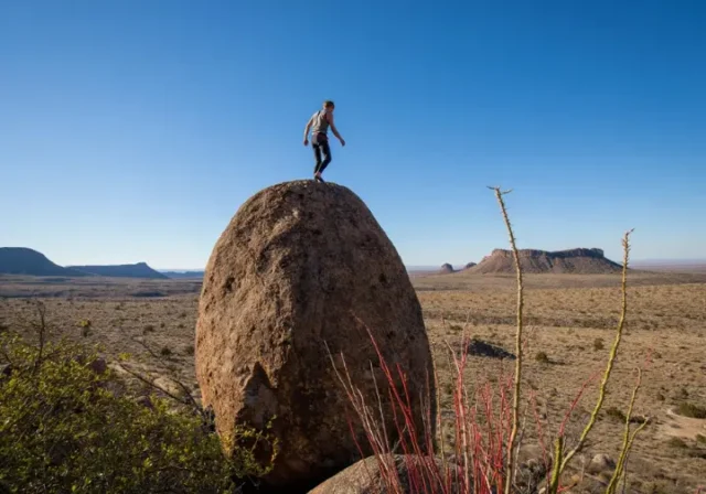 01 big-bend-bouldering-area-essential-tx-guide Climber successfully reaching the top of a large boulder in the Big Bend bouldering area, with the expansive Texas desert landscape in the background, illustrating an essential guide to bouldering in the region.