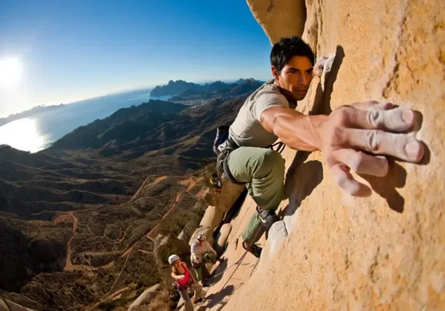 Two diverse climbers on a sunlit limestone cliff, showcasing various rock climbing styles available in Spain, with mountains and coastline in the background. Represents a definitive guide to rock climbing in Spain.