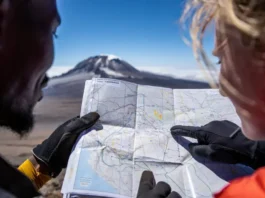 Two diverse climbers' hands holding a map of Kilimanjaro routes with Uhuru Peak in the background, symbolizing strategic planning for a mountain climbing expedition to Kilimanjaro.