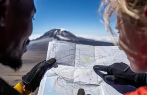 Mountain Climbing Kilimanjaro: The Summit Blueprint Two diverse climbers' hands holding a map of Kilimanjaro routes with Uhuru Peak in the background, symbolizing strategic planning for a mountain climbing expedition to Kilimanjaro.
