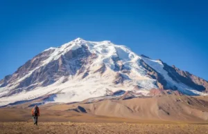 Mountaineering in Peru: Your Expert Ascent Guide Panoramic view of a towering snow-covered peak in the Peruvian Andes, with a tiny mountaineer figure in the distance, illustrating an expert guide to mountaineering in Peru.