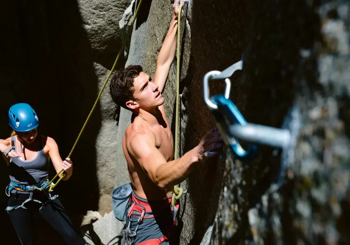 A climber demonstrating proper quickdraw clipping technique on a rock face, with a belayer in the background, showing mastery of quickdraw climbing.