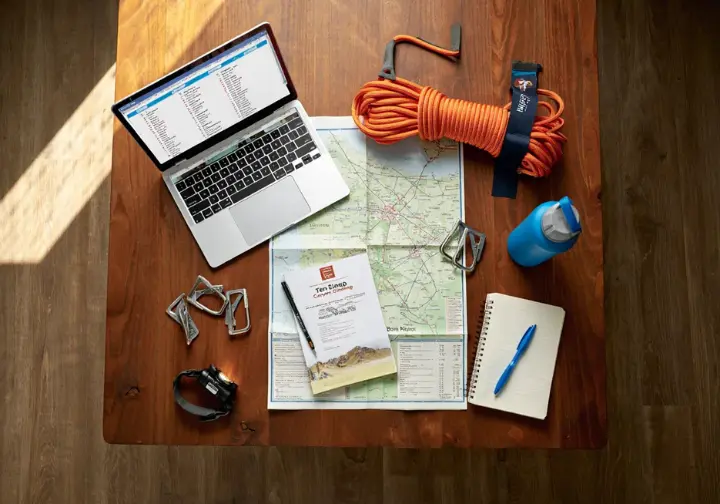 Organized flat lay of Ten Sleep rock climbing trip planning items: guidebook, map, laptop, and climbing gear, for logistics and preparation.