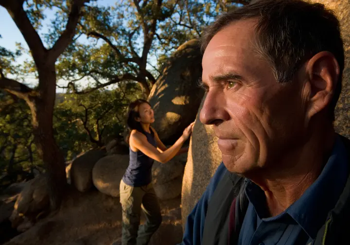 A seasoned climber thoughtfully observes the historic Boat Rock bouldering area as a younger climber studies a boulder, symbolizing its rich legacy and future preservation.