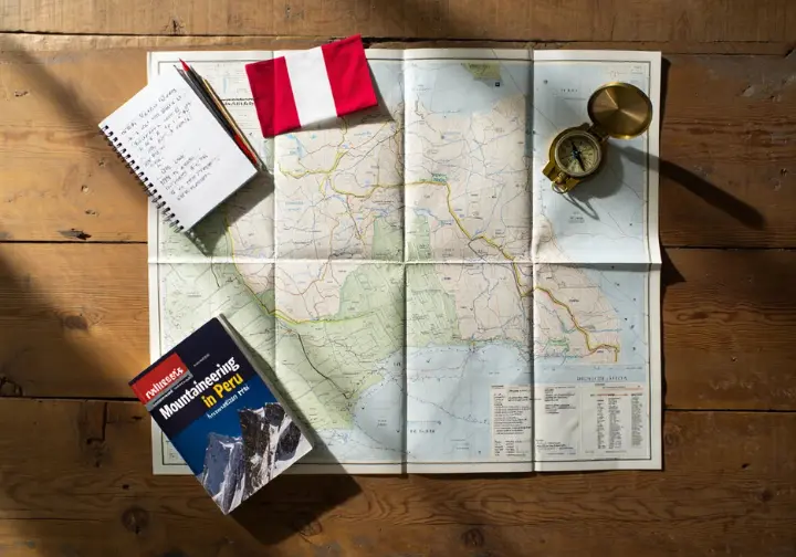 Flat lay of expedition planning tools like a map, compass, and guidebook for navigating logistics and regulations for mountaineering in Peru.