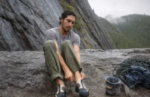 Why 98% of Climbers Buy Climbing Shoes Too Tight Climber sitting on a crag boulder easing on a Butora Endeavor climbing shoe before a route