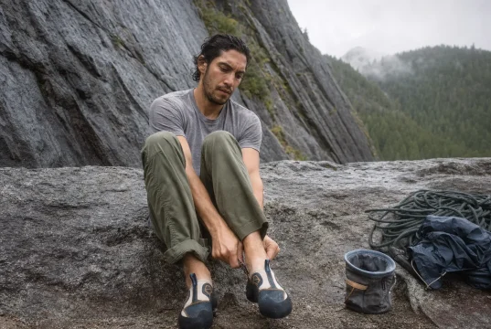 Why 98% of Climbers Buy Climbing Shoes Too Tight Climber sitting on a crag boulder easing on a Butora Endeavor climbing shoe before a route