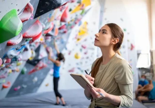 Climber thoughtfully analyzing a bouldering wall with a notebook, symbolizing a systematic approach to unlock potential and get better at bouldering.