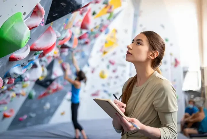 Climber thoughtfully analyzing a bouldering wall with a notebook, symbolizing a systematic approach to unlock potential and get better at bouldering.