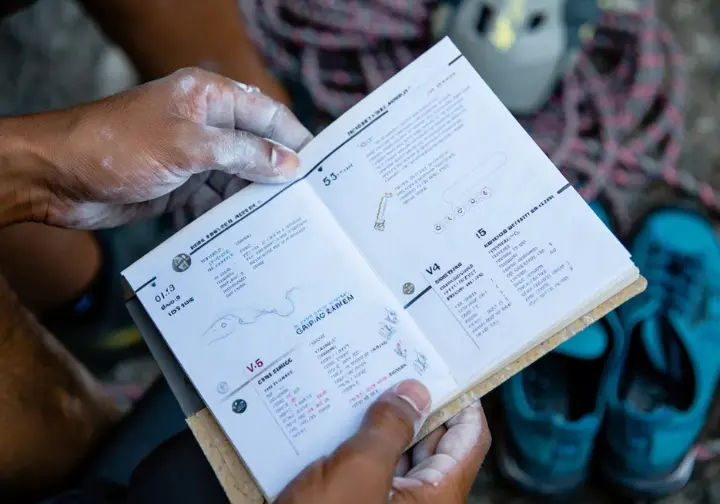 Diverse climbers' hands studying a guidebook showing different climbing grades, illustrating the system for understanding the difficulty of the world's hardest routes.