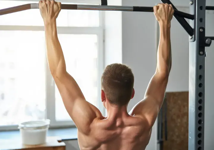 Person performing a dead hang, a foundational grip strength exercise for climbing, in a well-lit gym.