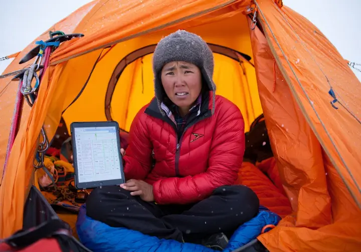 Budgeting for the unexpected on Everest: Climber reviewing budget on a tablet inside a tent during challenging conditions, needing a contingency fund.