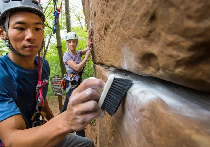 Climbers demonstrating responsible practices (brushing holds, safety check) for rock climbing in New River Gorge National Park.