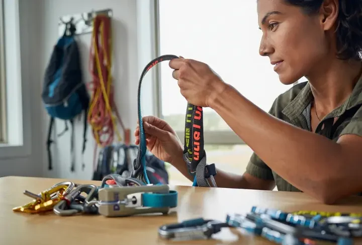 A female climber performing a crucial check by inspecting her climbing harness, with carabiners and other hardware laid out for maintenance.