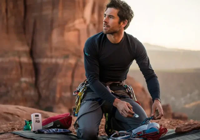 An experienced climber organizing next-level safety gear, including a PLB and first-aid kit, at the base of a cliff, illustrating essential climb safety beyond the basics.