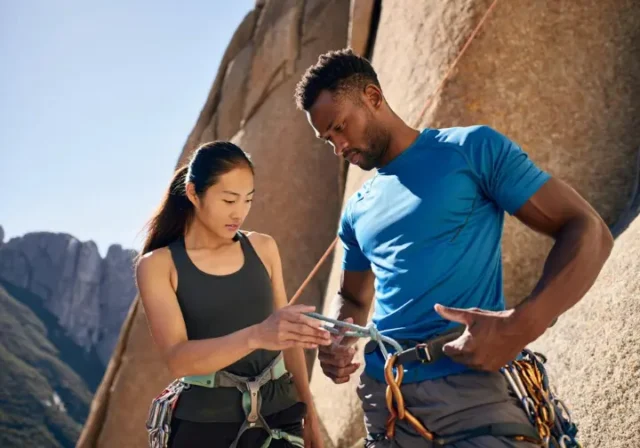 Two diverse climbers performing a pre-climb partner check at the base of a cliff, illustrating a key aspect of life-saving prevention from common climbing dangers.