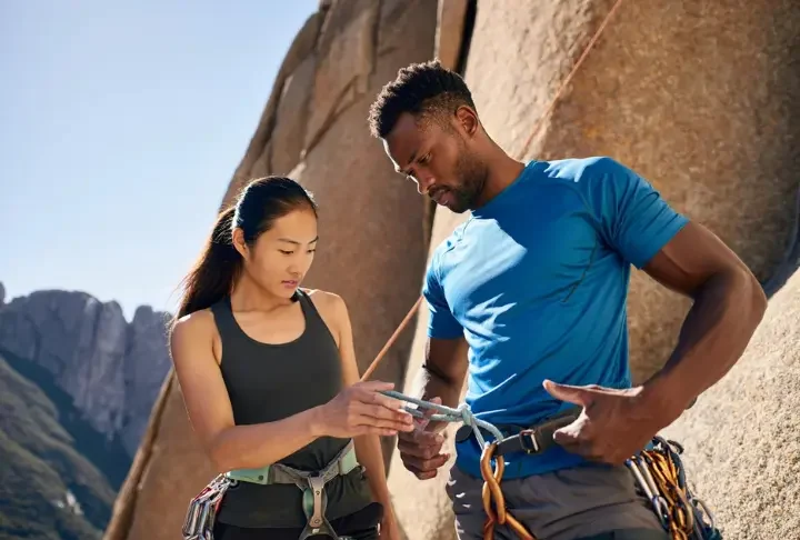 Two diverse climbers performing a pre-climb partner check at the base of a cliff, illustrating a key aspect of life-saving prevention from common climbing dangers.