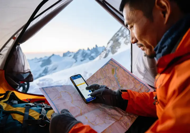 Alpinist inside a tent at a snowy base camp, studying a topo map and weather forecast, surrounded by essential gear, demonstrating preparedness for alpine climbing success.